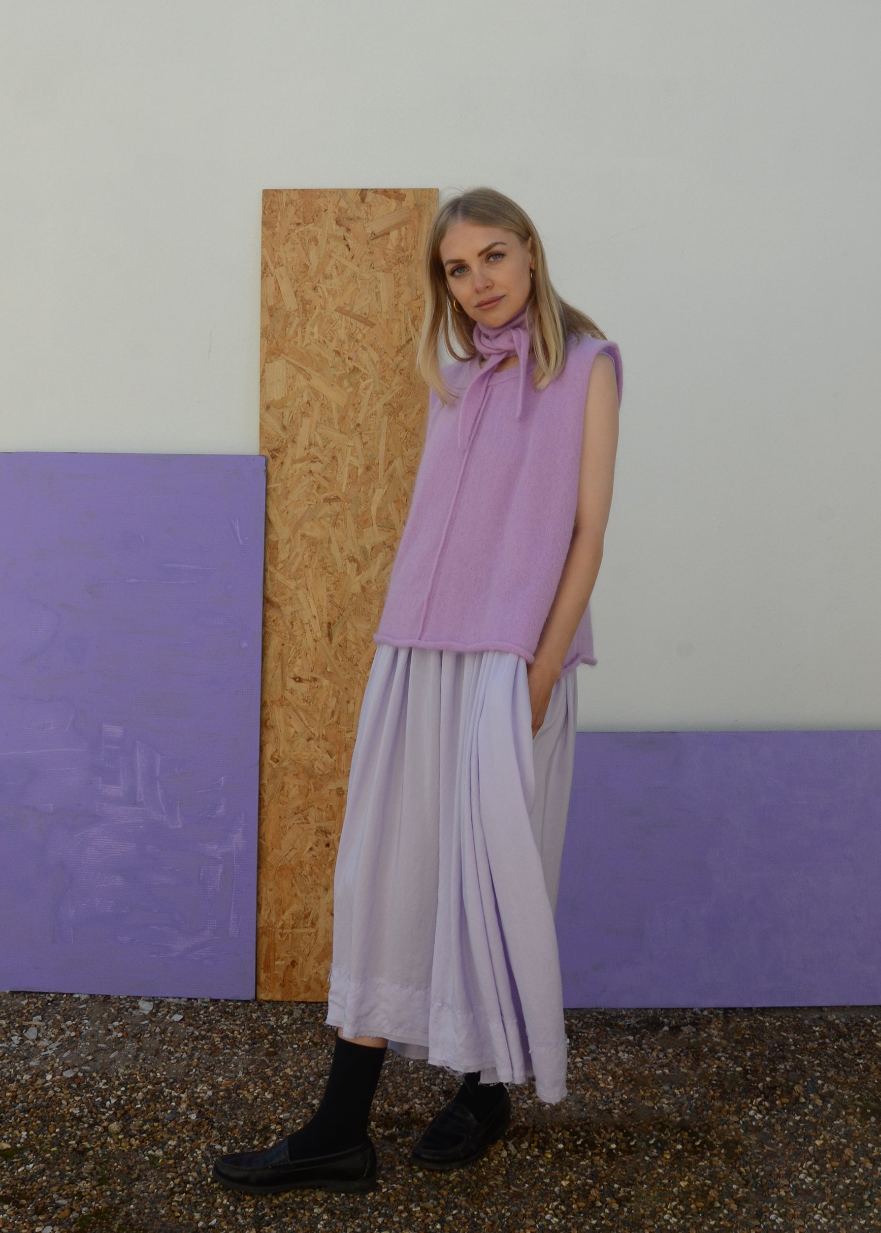 Woman wearing a light purple sleeveless top and pale lilac skirt standing against a textured wall with purple panels.